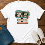 A white short-sleeve graphic T-shirt laid flat on a wooden surface, styled with folded blue jeans and white sneakers. The shirt features a retro marquee-style slogan print reading 'NOT UP FOR DEBATE' with additional text below.