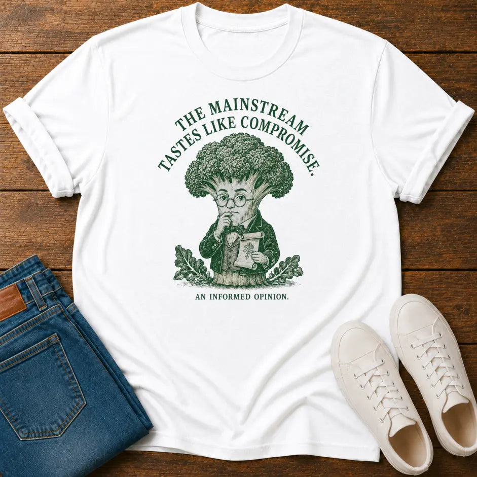 A white graphic T-shirt laid flat on a wooden surface, styled with folded blue jeans and white sneakers. The shirt has a green illustrated broccoli character and the text 'THE MAINSTREAM TASTES LIKE COMPROMISE.