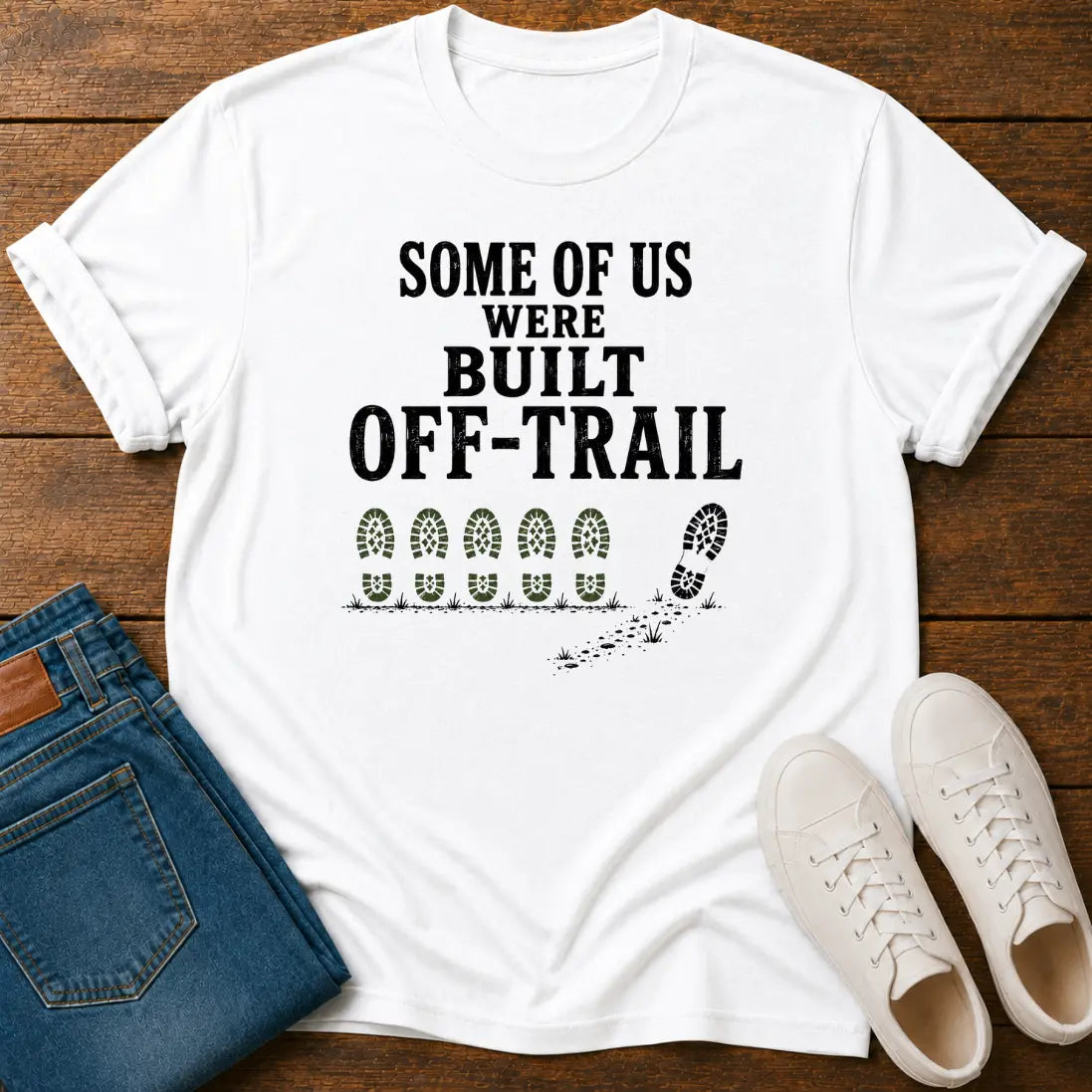 A white graphic T-shirt laid flat on a wooden surface, styled with folded blue jeans and white sneakers. The shirt has black text reading 'SOME OF US WERE BUILT OFF-TRAIL' above a row of bootprints and one larger bootprint with a trail effect.