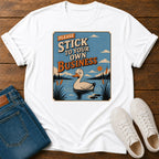 A white short-sleeve graphic T-shirt laid flat on a wooden surface, styled with folded blue jeans and white sneakers. The shirt features a large square vintage-style print showing a duck in a pond with the text 'PLEASE STICK TO YOUR OWN BUSINESS.