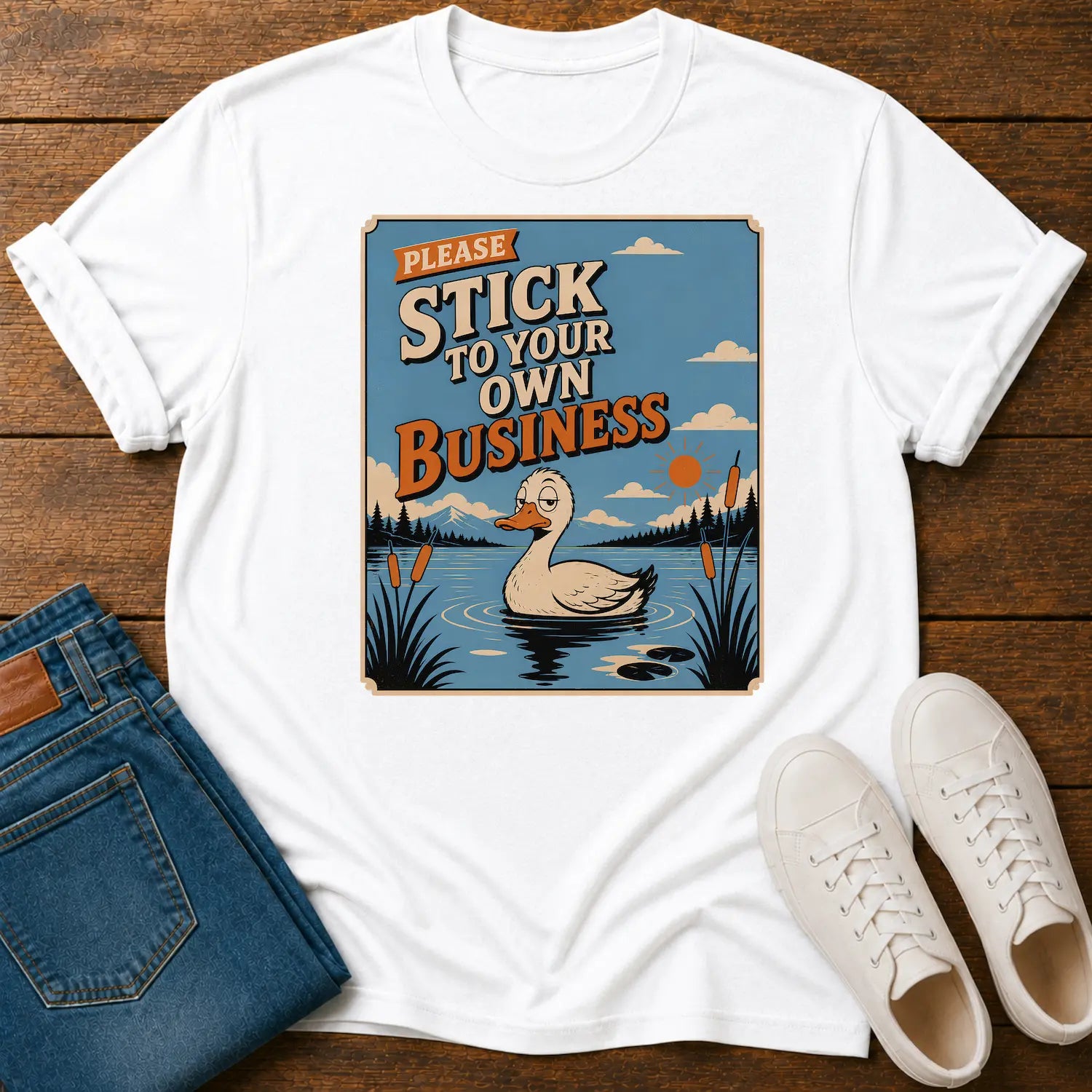A white short-sleeve graphic T-shirt laid flat on a wooden surface, styled with folded blue jeans and white sneakers. The shirt features a large square vintage-style print showing a duck in a pond with the text 'PLEASE STICK TO YOUR OWN BUSINESS.