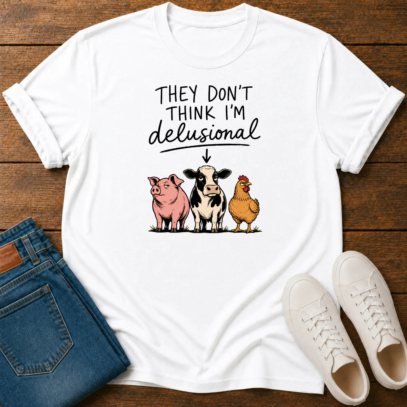 A white graphic t-shirt laid flat on a dark wood background, styled with blue jeans and white sneakers. The t-shirt features the text 'THEY DON'T THINK I'M delusional' above an illustration of a pink pig, a black-and-white cow, and a yelloe chicken.