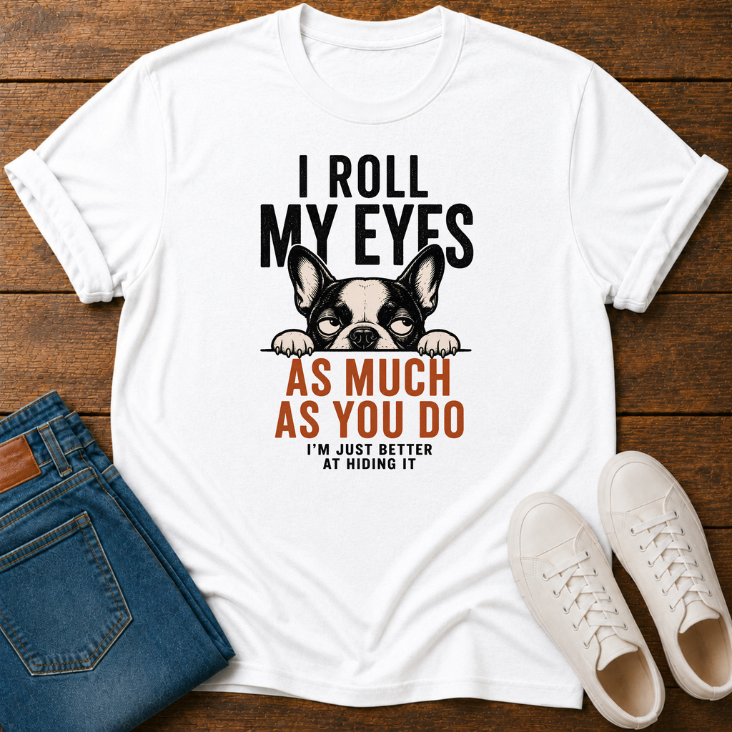A white graphic t-shirt laid flat on a dark wood background, styled with folded blue jeans and white sneakers. The t-shirt features a black and white French Bulldog graphic with the text 'I ROLL MY EYES AS MUCH AS YOU DO I'M JUST BETTER AT HIDING IT' in black and orange lettering.
