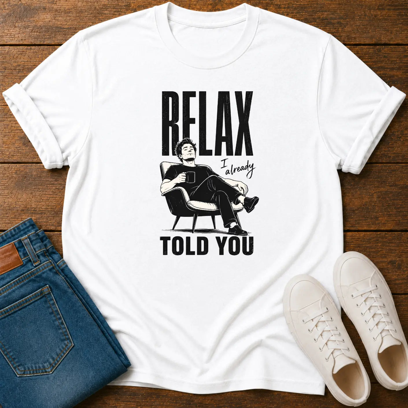 A white graphic t-shirt laid flat on a dark wood background, styled with folded blue jeans and a pair of white sneakers. The t-shirt features a black graphic of a man sitting in a chair with the text 'RELAX I already TOLD YOU'.