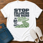 A white graphic t-shirt laid flat on a wooden surface, styled with folded blue jeans and white sneakers. The t-shirt features a graphic that reads 'STOP FOLLOWING THE HERD' and 'CHOOSE THE GREEN PATH' with an illustration of a cow and a green leafy path.