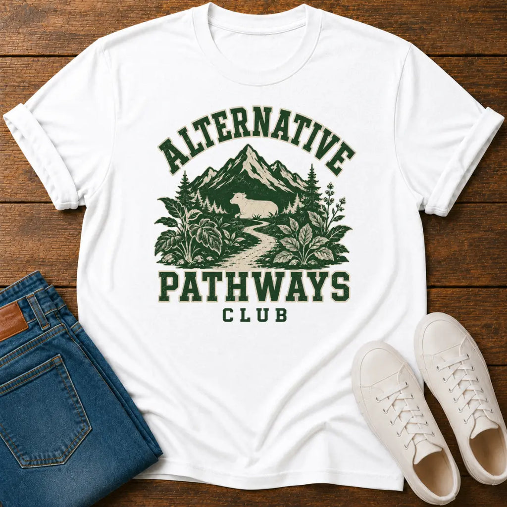 A white short-sleeve graphic T-shirt laid flat on a wooden surface, styled with folded blue jeans and white sneakers. The shirt has a dark green outdoor-style print reading “ALTERNATIVE PATHWAYS CLUB” with mountains, trees, and a path graphic.
