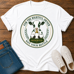 A white graphic t-shirt laid flat on a wooden surface, styled with blue jeans and white sneakers. The t-shirt features a circular graphic of a black and white cow holding a gold bell, with the text 'FOR THE RIGHTFUL OWNER' and 'NOT YOUR BOTTLE' in green and gold.