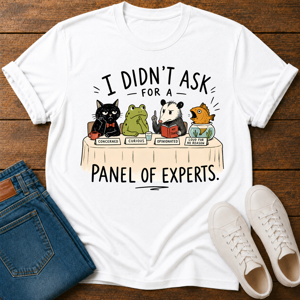 A white graphic t-shirt laid flat on a dark wood background, styled with blue jeans and white sneakers. The t-shirt features a cartoon panel discussion graphic of a black cat, green frog, panda, and fish, with the text 'I DIDN'T ASK FOR A PANEL OF EXPERTS.