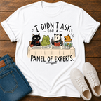 A white graphic t-shirt laid flat on a dark wood background, styled with blue jeans and white sneakers. The t-shirt features a cartoon panel discussion graphic of a black cat, green frog, panda, and fish, with the text 'I DIDN'T ASK FOR A PANEL OF EXPERTS.