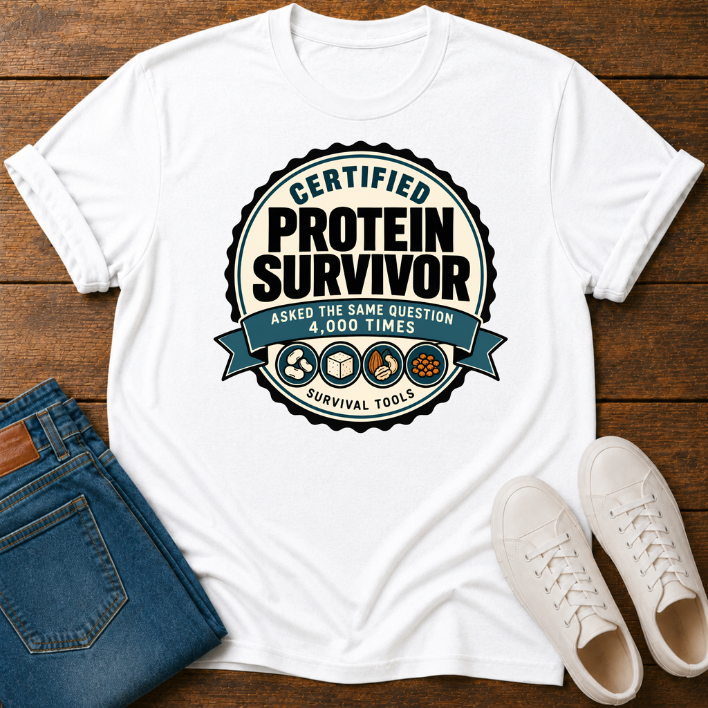 A white short-sleeve graphic T-shirt laid flat on a wooden surface, styled with folded blue jeans and white sneakers. The shirt features a large circular badge design reading 'Certified Protein Survivor' with additional text and small survival-themed icons.