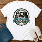 A white short-sleeve graphic T-shirt laid flat on a wooden surface, styled with folded blue jeans and white sneakers. The shirt features a large circular badge design reading 'Certified Protein Survivor' with additional text and small survival-themed icons.