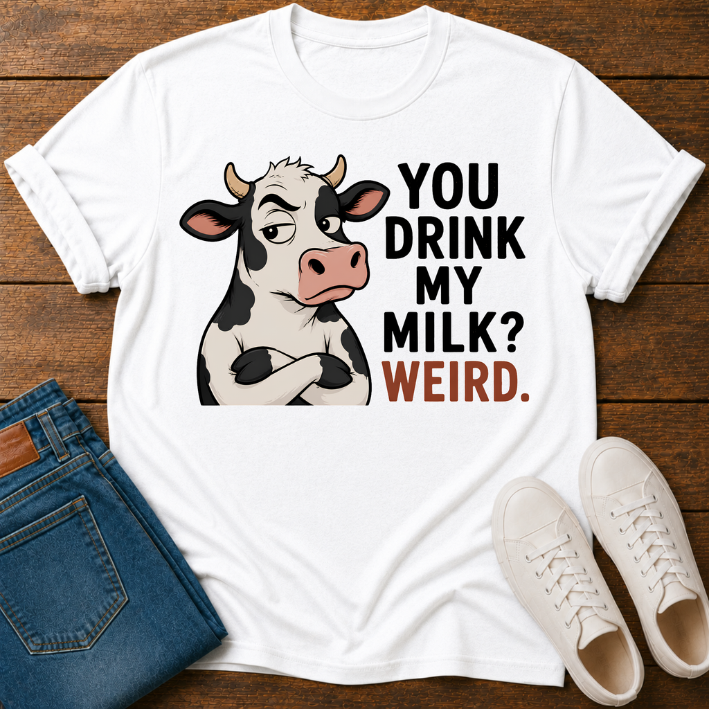 A white graphic t-shirt laid flat on a dark wood background, styled with folded blue jeans and white sneakers. The t-shirt features a cartoon cow with crossed arms next to the text 'YOU DRINK MY MILK? WEIRD.