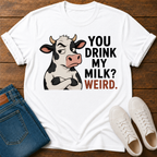 A white graphic t-shirt laid flat on a dark wood background, styled with folded blue jeans and white sneakers. The t-shirt features a cartoon cow with crossed arms next to the text 'YOU DRINK MY MILK? WEIRD.