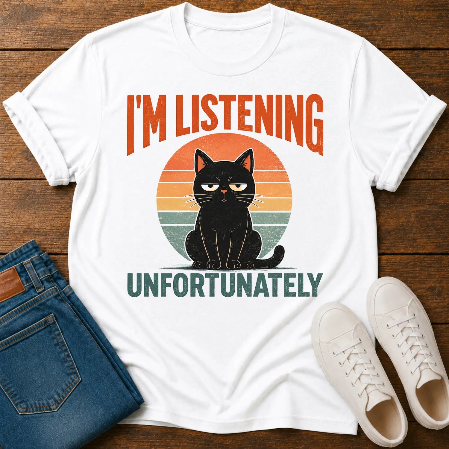 A white graphic t-shirt laid flat on a wooden surface, styled with folded blue jeans and a pair of white sneakers. The t-shirt features a retro-style graphic of a black cat sitting in front of a sunset circle, with the text 'I'M LISTENING UNFORTUNATELY' in orange and dark green.